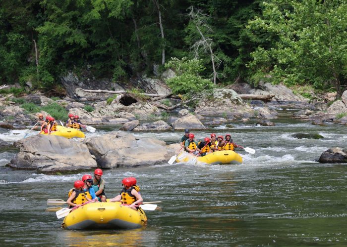 Asheville tourists in yellow rafts smiling after completing a run of rapids on FB9 surrounded by old volcanic rock and lush green forests in the Blue Ridge Mountains.